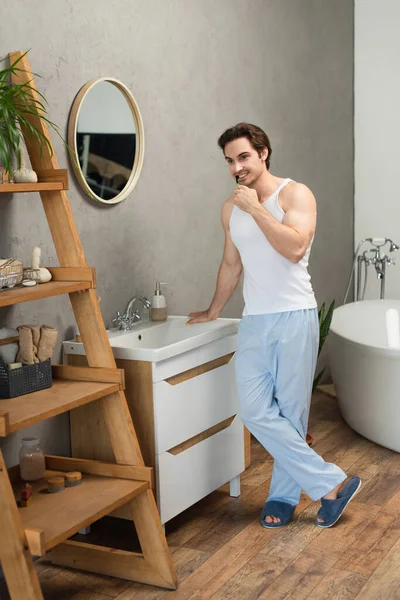 Homem em top tanque branco e calças de pijama azul em pé perto de pia no banheiro e escovação de dentes — Fotografia de Stock