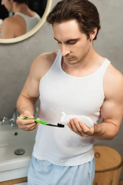 Morena hombre en camiseta blanca aplicando pasta de dientes en el cepillo de dientes en el baño - foto de stock