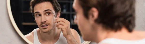 Young man tweezing eyebrows near mirror on blurred foreground, banner — Stock Photo