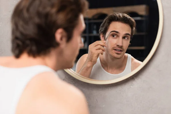 Brunette man tweezing eyebrows while looking in mirror — Stock Photo