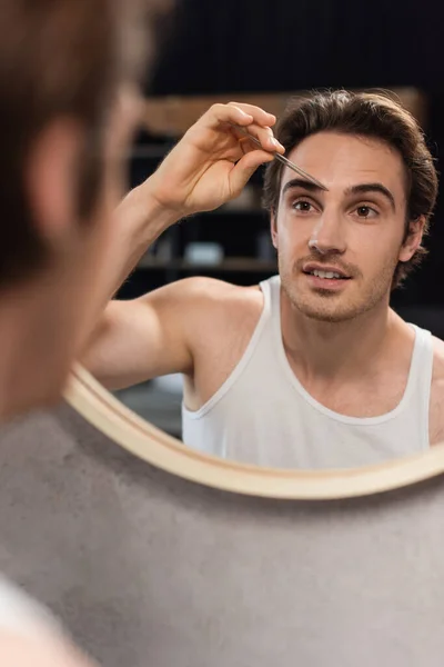 Man in white tank top tweezing eyebrows near mirror — Stock Photo