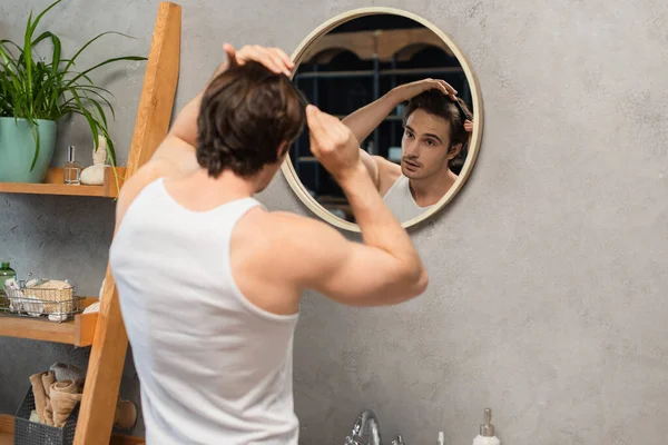 Jeune homme regardant dans le miroir tout en peignant les cheveux dans la salle de bain — Photo de stock