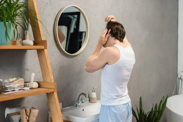 Uomo in canotta bianca pettinatura capelli vicino specchio e lavandino in bagno — Foto stock