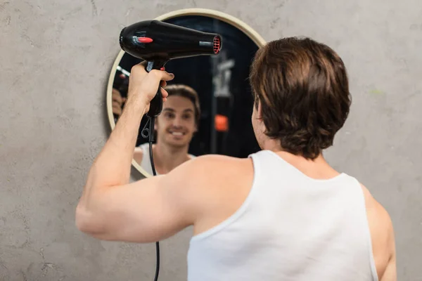 Glücklicher junger Mann trocknet Haare, während er im Badezimmer in den Spiegel schaut — Stockfoto