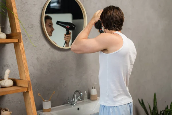 Homme en blanc débardeur sèche-cheveux près du miroir dans la salle de bain — Photo de stock