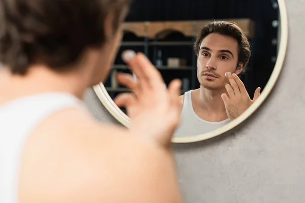 Jeune homme appliquant crème visage près du miroir dans la salle de bain — Photo de stock