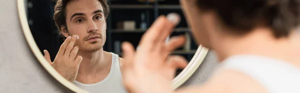 Uomo guardando nello specchio del bagno e applicando crema per il viso, banner — Foto stock