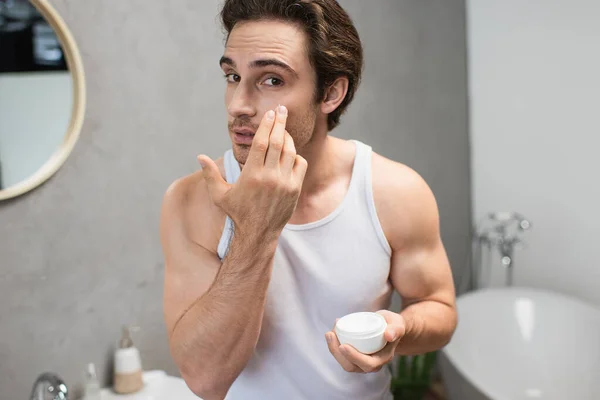 Hombre en camiseta blanca mirando a la cámara mientras se aplica crema facial - foto de stock