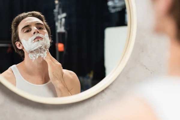 Uomo bruna applicare schiuma da barba sul viso mentre si guarda allo specchio — Foto stock