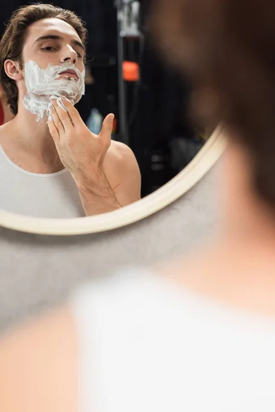 Giovane uomo che applica schiuma da barba sul viso vicino specchio in bagno in primo piano sfocato — Foto stock