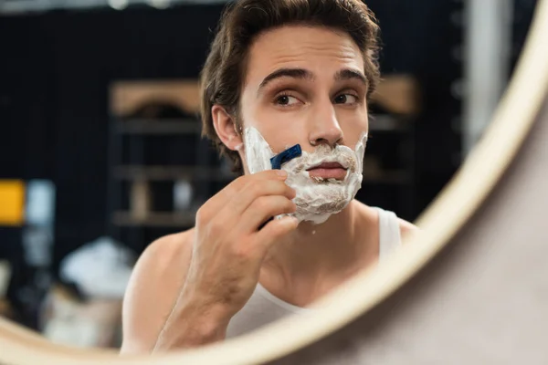 Joven morena hombre mirando en espejo mientras afeitarse en la mañana - foto de stock