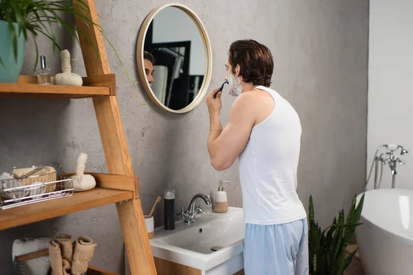 Joven en blanco tanque superior afeitado cerca de estante de madera y espejo en el baño - foto de stock