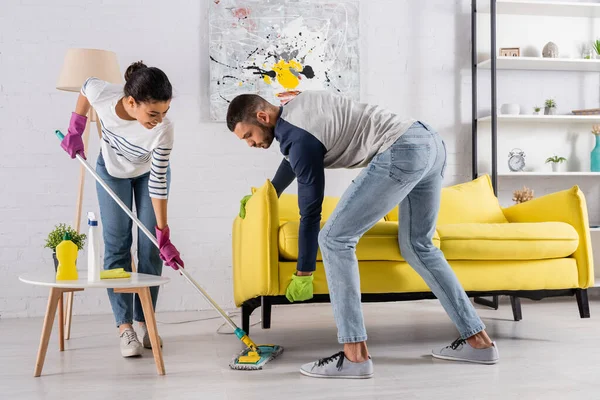 Jovem casal interracial limpeza chão perto do sofá em casa — Fotografia de Stock