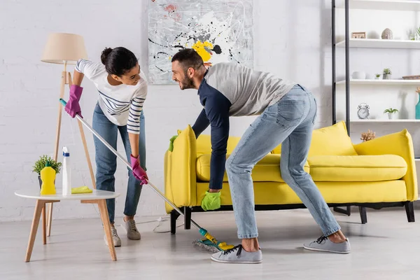Allegro multietnico coppia pulizia pavimento sotto il divano a casa — Foto stock