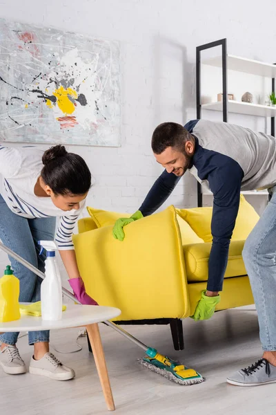 Smiling man lifting couch near african american girlfriend with mop at home — Stock Photo