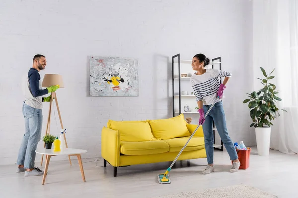 Side view of cheerful multiethnic couple with mop and rag cleaning living room — Stock Photo