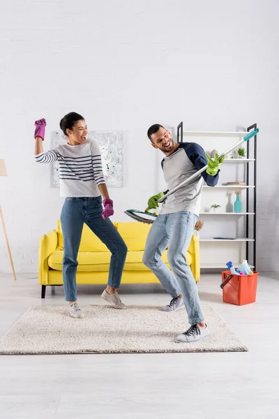 Joyeux couple interracial en gants en caoutchouc jouant avec la serpillière — Photo de stock