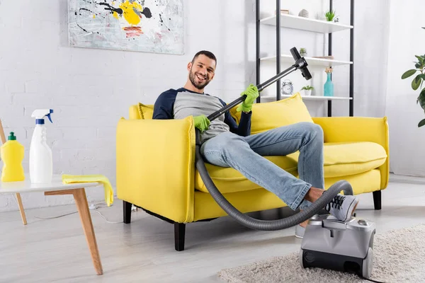 Smiling man holding vacuum cleaner while sitting on couch at home — Stock Photo