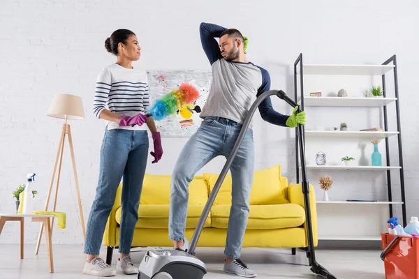 Femme afro-américaine avec brosse à poussière regardant petit ami avec aspirateur grimacing à la maison — Photo de stock