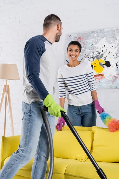 Sorridente donna afroamericana con spazzola antipolvere guardando fidanzato con aspirapolvere — Foto stock