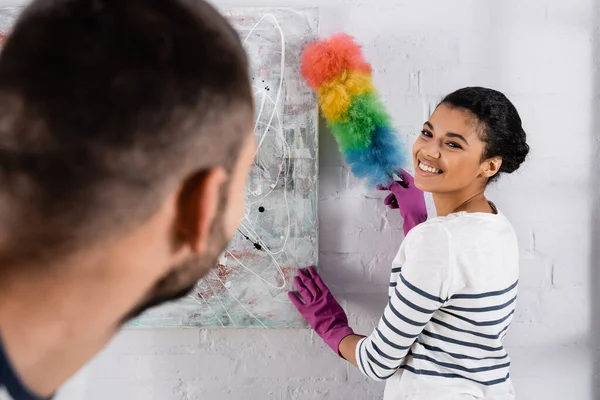 Smiling african american woman cleaning drawing with dust brush near blurred boyfriend — Stock Photo