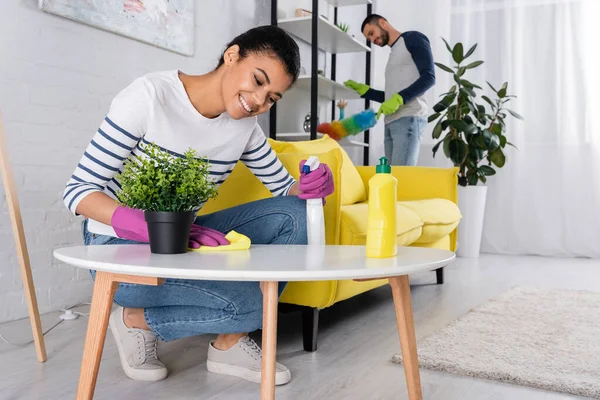 Positive african american woman cleaning coffee table near blurred boyfriend — Stock Photo