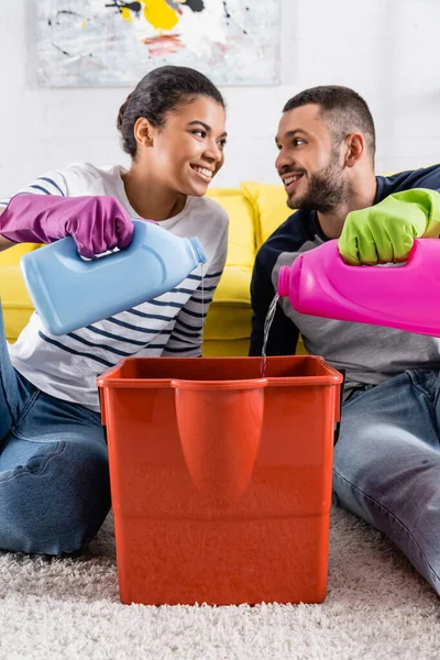 Sonriente pareja interracial vertiendo detergentes en cubo - foto de stock