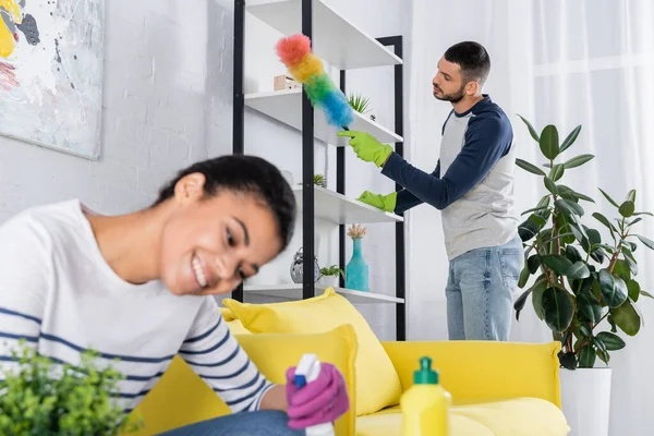Jovem armário de limpeza com escova de poeira perto da namorada afro-americana borrada — Fotografia de Stock