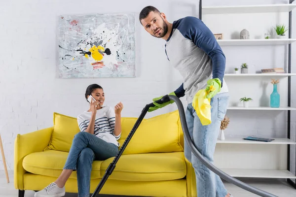 Confused man with rag and vacuum cleaner standing near african american girlfriend talking on smartphone — Stock Photo