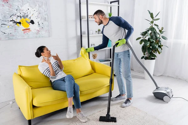 Side view of angry man with rag and vacuum cleaner looking at african american girlfriend with smartphone on couch — Stock Photo