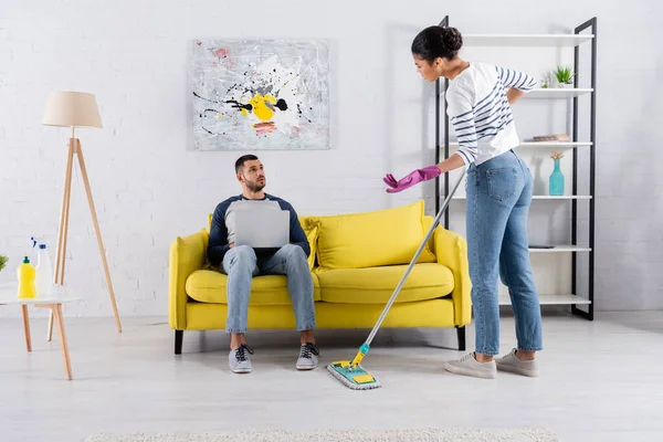 Femme afro-américaine en colère avec serpillière regardant petit ami avec ordinateur portable sur le canapé — Photo de stock