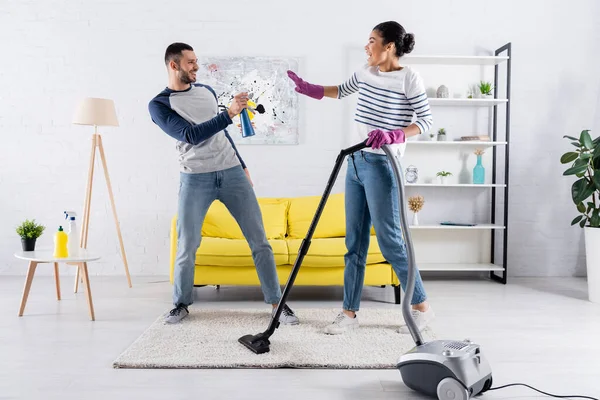 Vista lateral del hombre alegre sosteniendo detergente cerca de la novia afroamericana con aspiradora - foto de stock