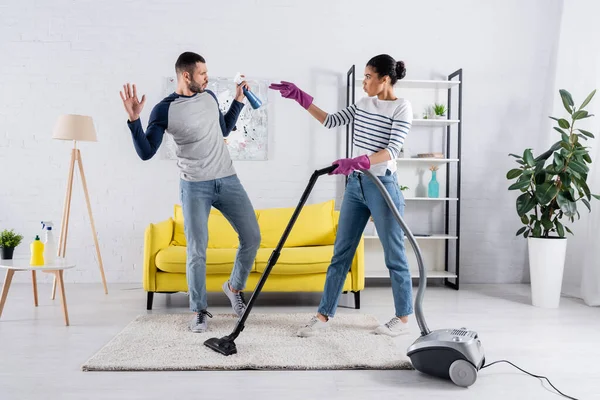 Side view of interracial couple with detergent and vacuum cleaner having fun in living room — Stock Photo