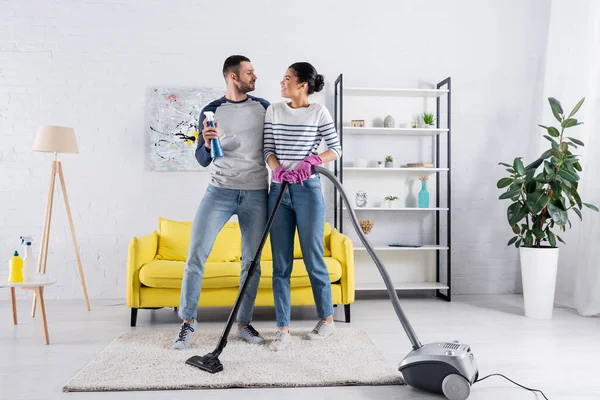 Vue latérale du couple interracial souriant avec des fournitures de nettoyage à la maison — Photo de stock