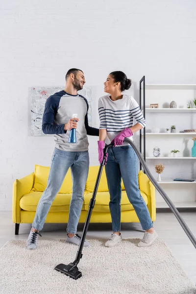 Vista lateral del hombre alegre sosteniendo detergente cerca de la novia afroamericana con aspiradora - foto de stock