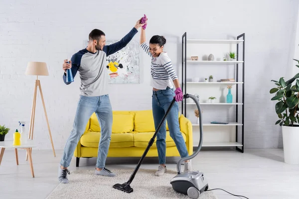 Feliz pareja multiétnica con suministros de limpieza bailando en el salón - foto de stock