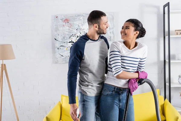 Couple interracial souriant avec aspirateur et détergent à la maison — Photo de stock