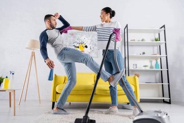 Side view of african american woman with vacuum cleaner pointing at boyfriend with detergent — Stock Photo