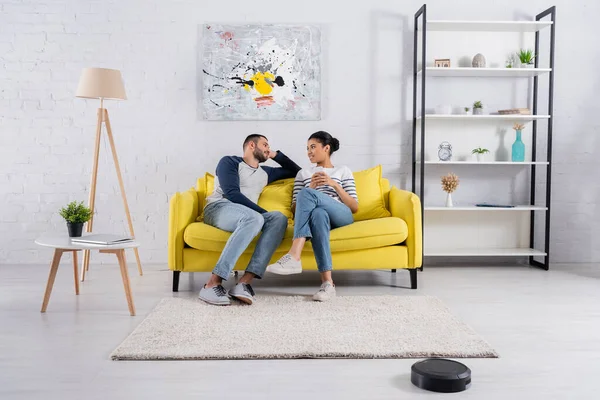 Joven pareja interracial con smartphone hablando cerca de la aspiradora robótica - foto de stock