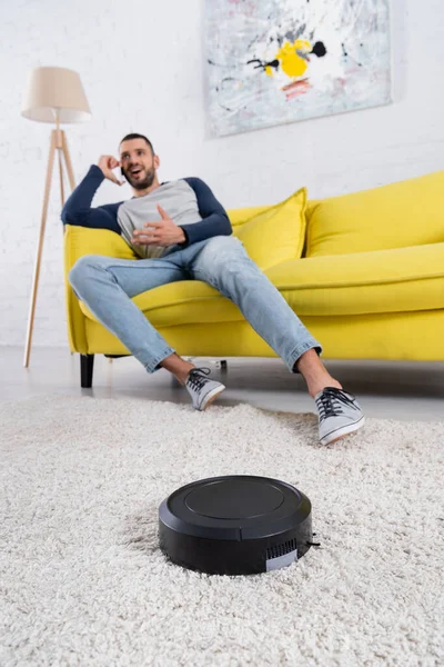 Robotic vacuum cleaner near blurred man talking on smartphone in living room — Stock Photo