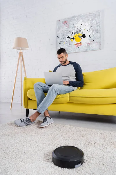 Young man using laptop near robotic vacuum cleaner in living room — Stock Photo