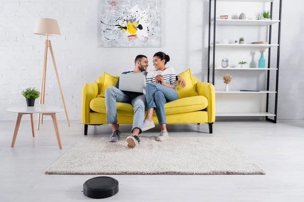 Couple souriant multiethnique utilisant des appareils près de l'aspirateur robotique à la maison — Photo de stock