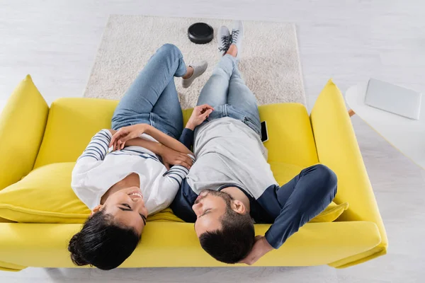 Vista superior de la sonriente pareja multiétnica sentada en el sofá cerca de la borrosa aspiradora robótica en la alfombra - foto de stock