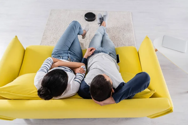 Вид сверху на многонациональную пару, сидящую рядом с устройствами и пылесосом на диване — стоковое фото