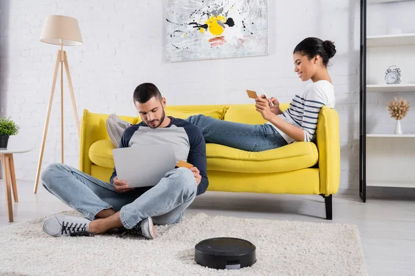 Smiling multiethnic couple using devices and credit cards near robotic vacuum cleaner — Stock Photo