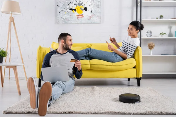 Pareja interracial con gadgets y tarjetas de crédito cerca de aspiradora robótica en casa — Stock Photo