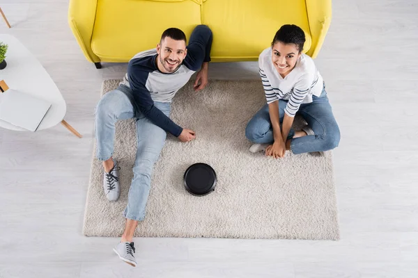 Vista aérea de una pareja interracial sonriente mirando la cámara cerca de la aspiradora robótica en la alfombra - foto de stock