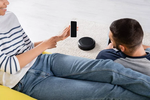 Smiling african american woman pointing at smartphone near boyfriend and vacuum cleaner at home — Stock Photo