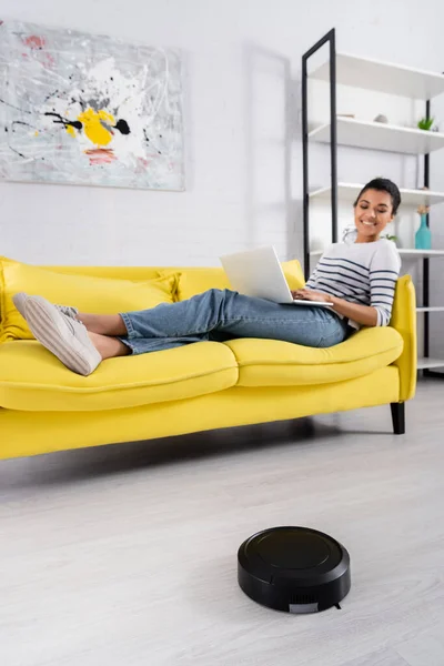 Robotic vacuum cleaner on floor near african american woman with laptop on blurred background — Stock Photo