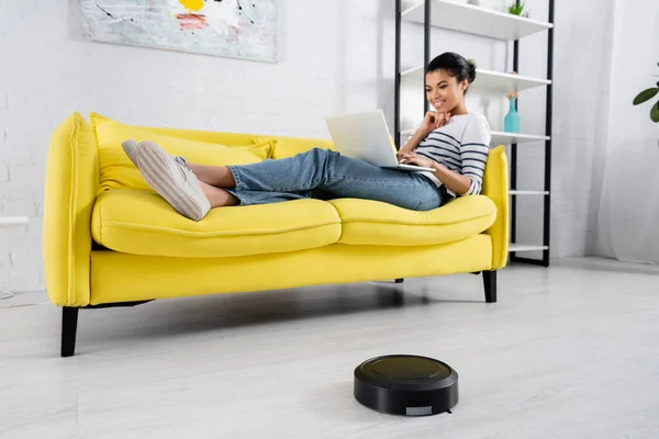 Robotic vacuum cleaner near african american woman using laptop in living room — Stock Photo